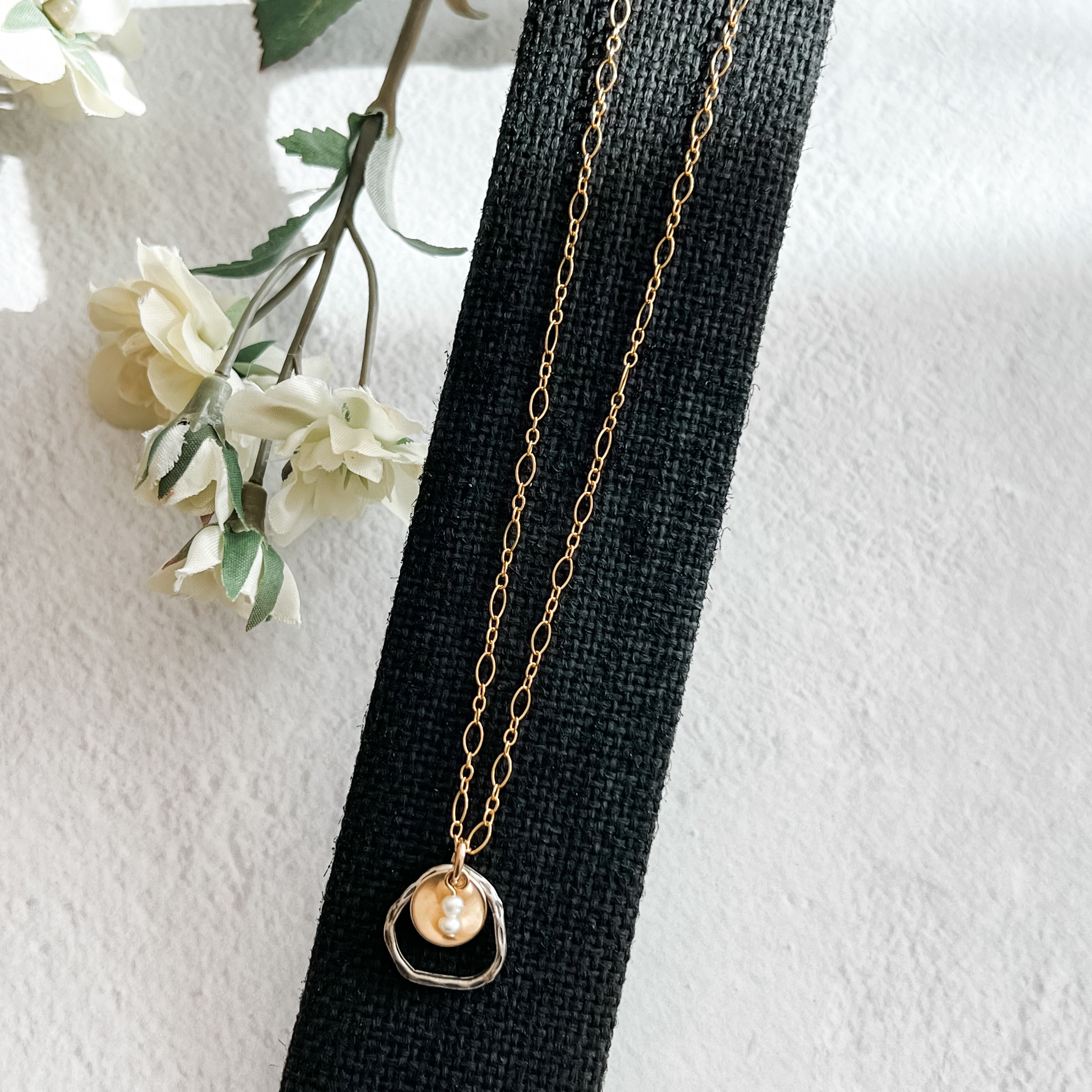 gold fill metal disc cupping two freshwater pearls with sterling open link on gold fill chain on black background with flowers to one side.