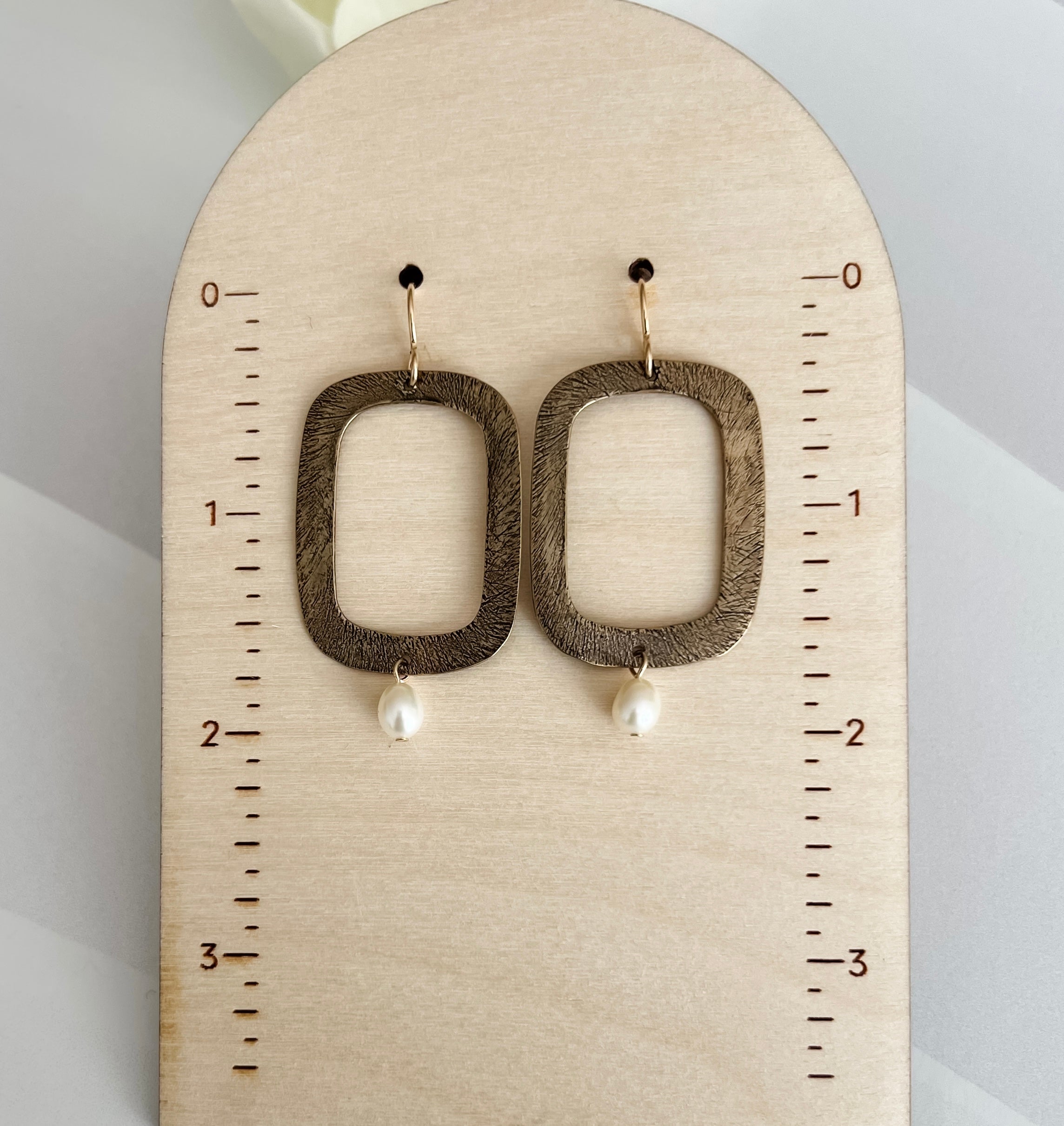 Handcrafted bronze rectangular hoop earrings with white freshwater pearl drops mounted on wooden measurement display, showing earrings measure approximately 2 inches in total length