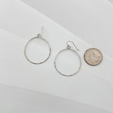 Handcrafted sterling silver hammered circle hoop earrings displayed beside a US quarter coin, showing approximately 1.5 inch diameter, lightweight everyday hoops