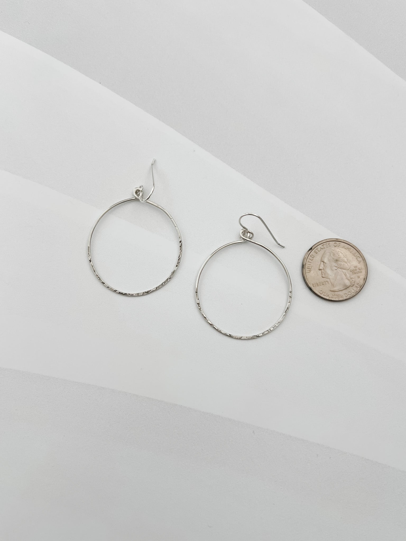 Handcrafted sterling silver hammered circle hoop earrings displayed beside a US quarter coin, showing approximately 1.5 inch diameter, lightweight everyday hoops