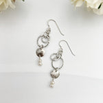 Caroline handcrafted sterling silver drop earrings with hammered texture, freshwater pearls, and interlocking circles on white background with flowers by Dawn Carlson Designs