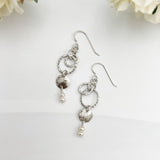Caroline handcrafted sterling silver drop earrings with hammered texture, freshwater pearls, and interlocking circles on white background with flowers by Dawn Carlson Designs