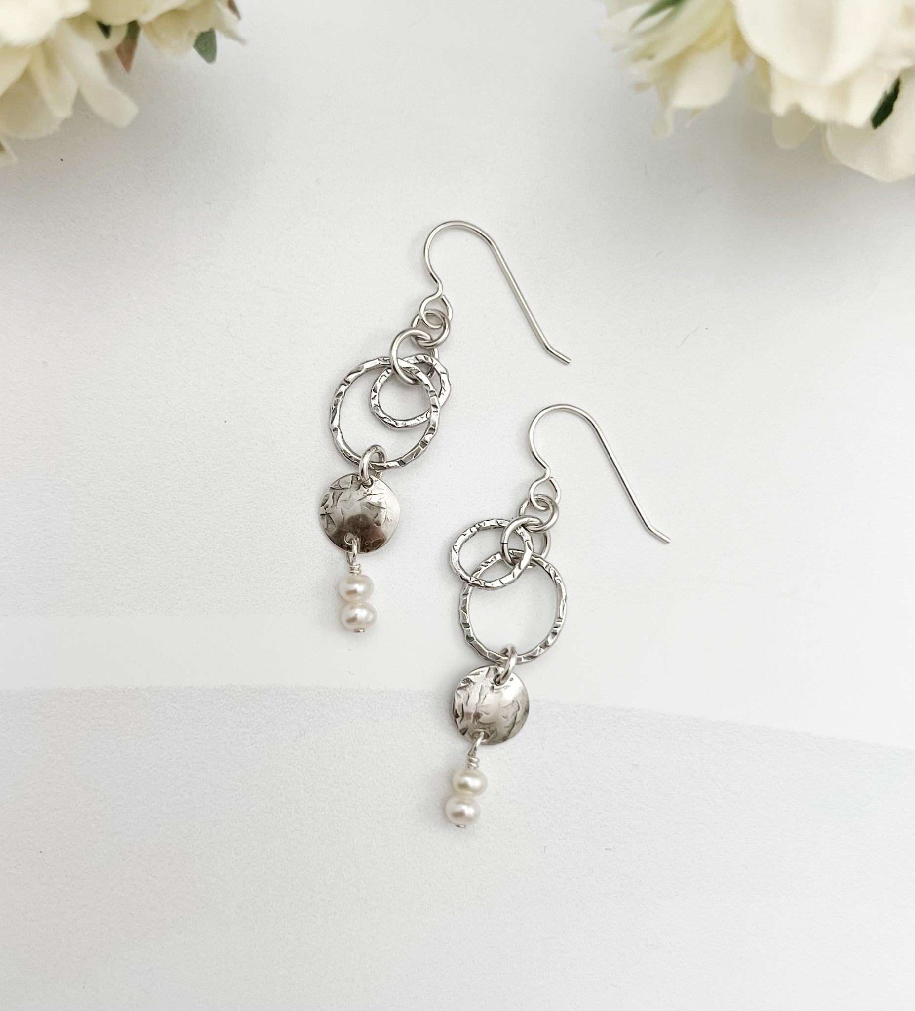 Caroline handcrafted sterling silver drop earrings with hammered texture, freshwater pearls, and interlocking circles on white background with flowers by Dawn Carlson Designs