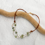 Double-strand freshwater pearl and leather necklace with green Czech glass beads, draped over driftwood on a white textured backgroun