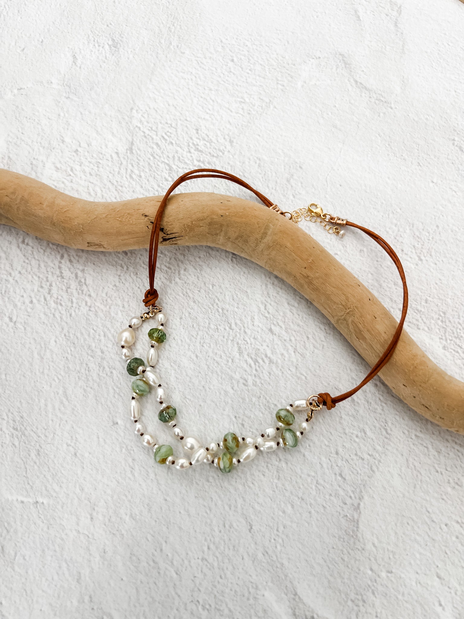 Double-strand freshwater pearl and leather necklace with green Czech glass beads, draped over driftwood on a white textured backgroun