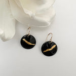 Handcrafted black enamel disc earrings with diagonal textured 24k gold foil accent on 14k gold fill French wires, by Dawn Carlson Designs. Displayed against a white background with a white fabric flower prop.