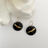 Handcrafted black enamel disc earrings with diagonal textured 24k gold foil accent on 14k gold fill French wires, by Dawn Carlson Designs. Displayed against a white background with a white fabric flower prop.