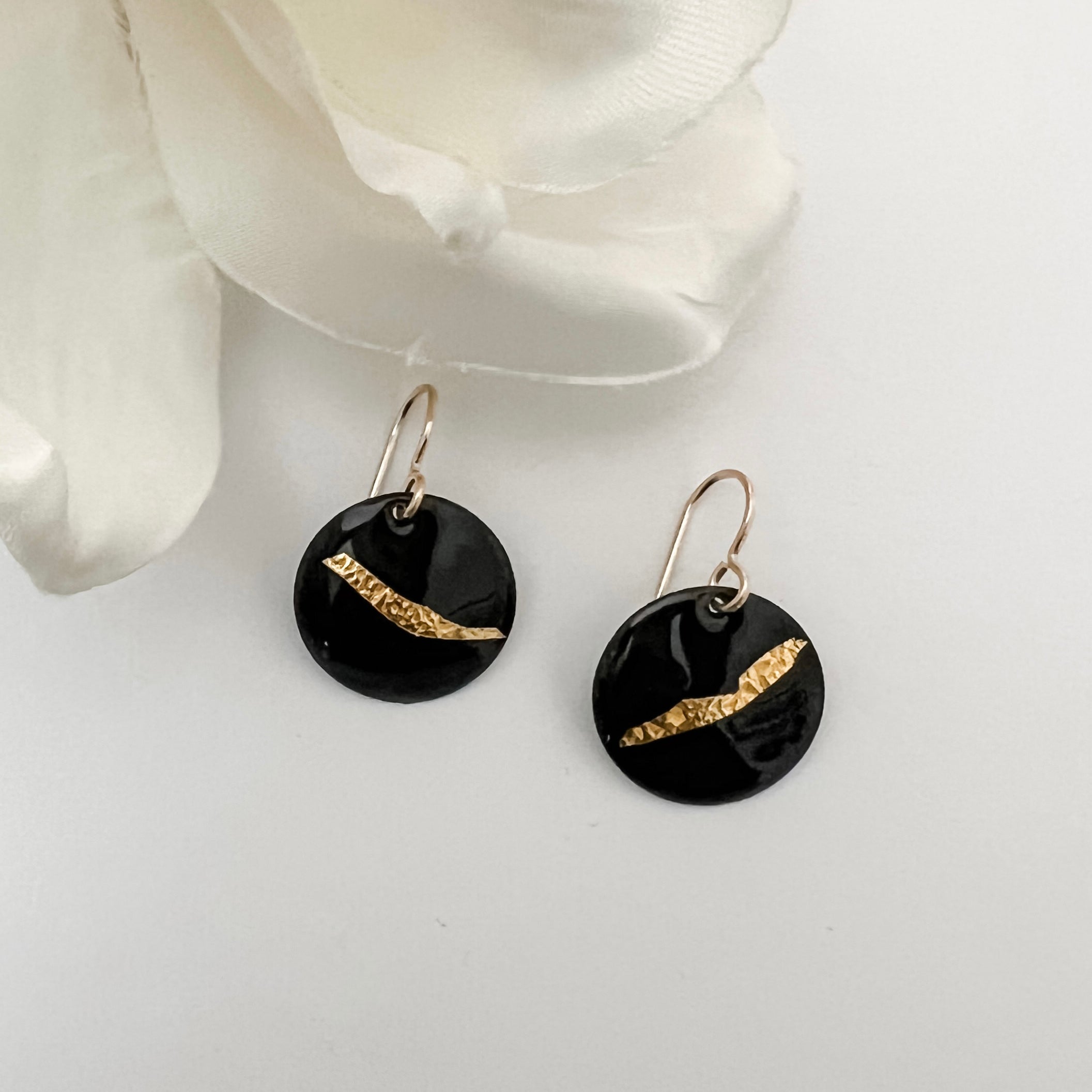 Handcrafted black enamel disc earrings with diagonal textured 24k gold foil accent on 14k gold fill French wires, by Dawn Carlson Designs. Displayed against a white background with a white fabric flower prop.