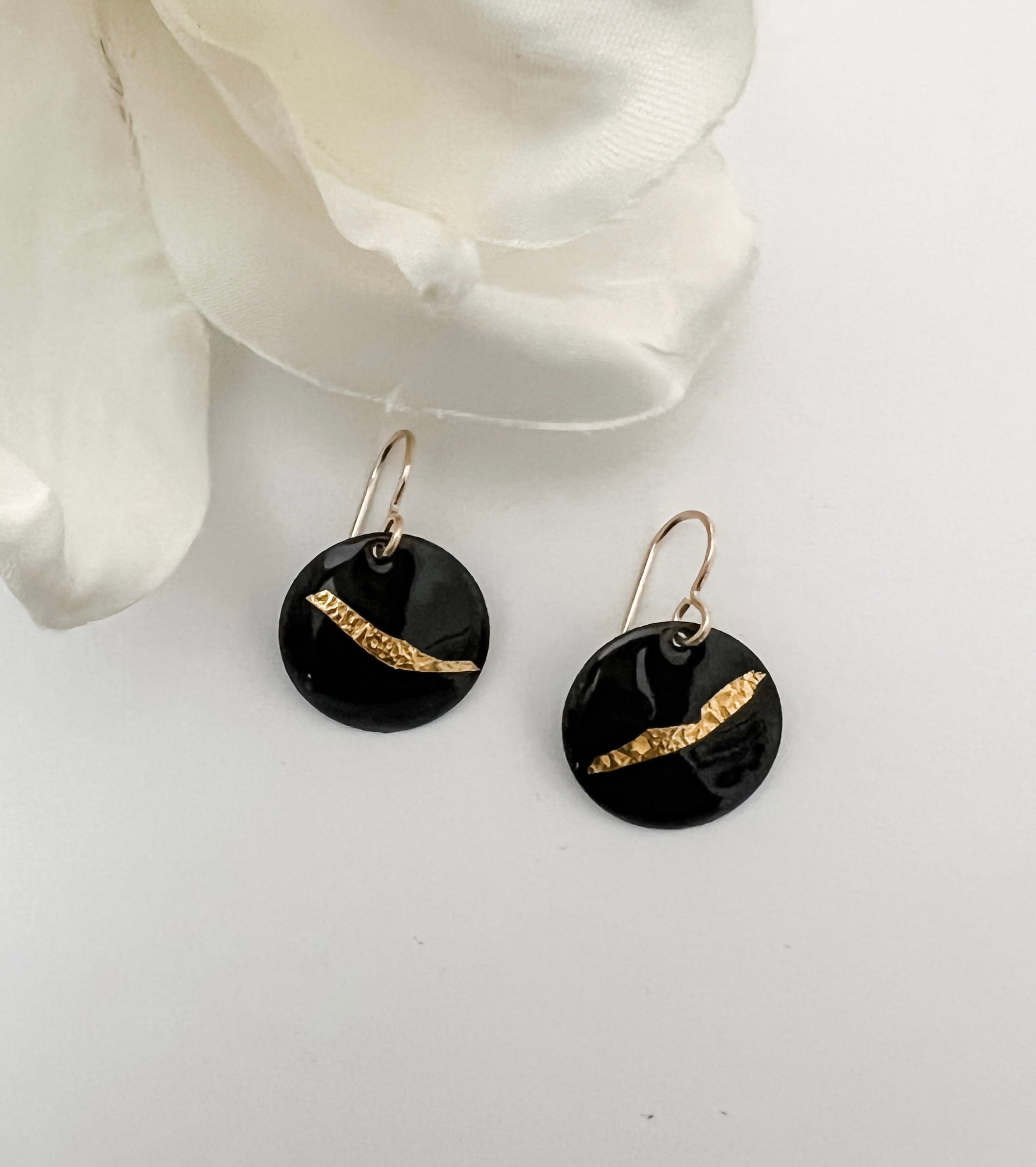 Handcrafted black enamel disc earrings with diagonal textured 24k gold foil accent on 14k gold fill French wires, by Dawn Carlson Designs. Displayed against a white background with a white fabric flower prop.