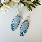 Handcrafted light blue enamel earrings with dark blue, cream, and seafoam splashes on sterling silver ear wires by Dawn Carlson Designs. Shown with fabric flowers.