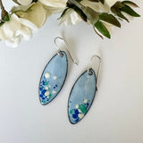 Handcrafted light blue enamel earrings with dark blue, cream, and seafoam splashes on sterling silver ear wires by Dawn Carlson Designs. Shown with fabric flowers.