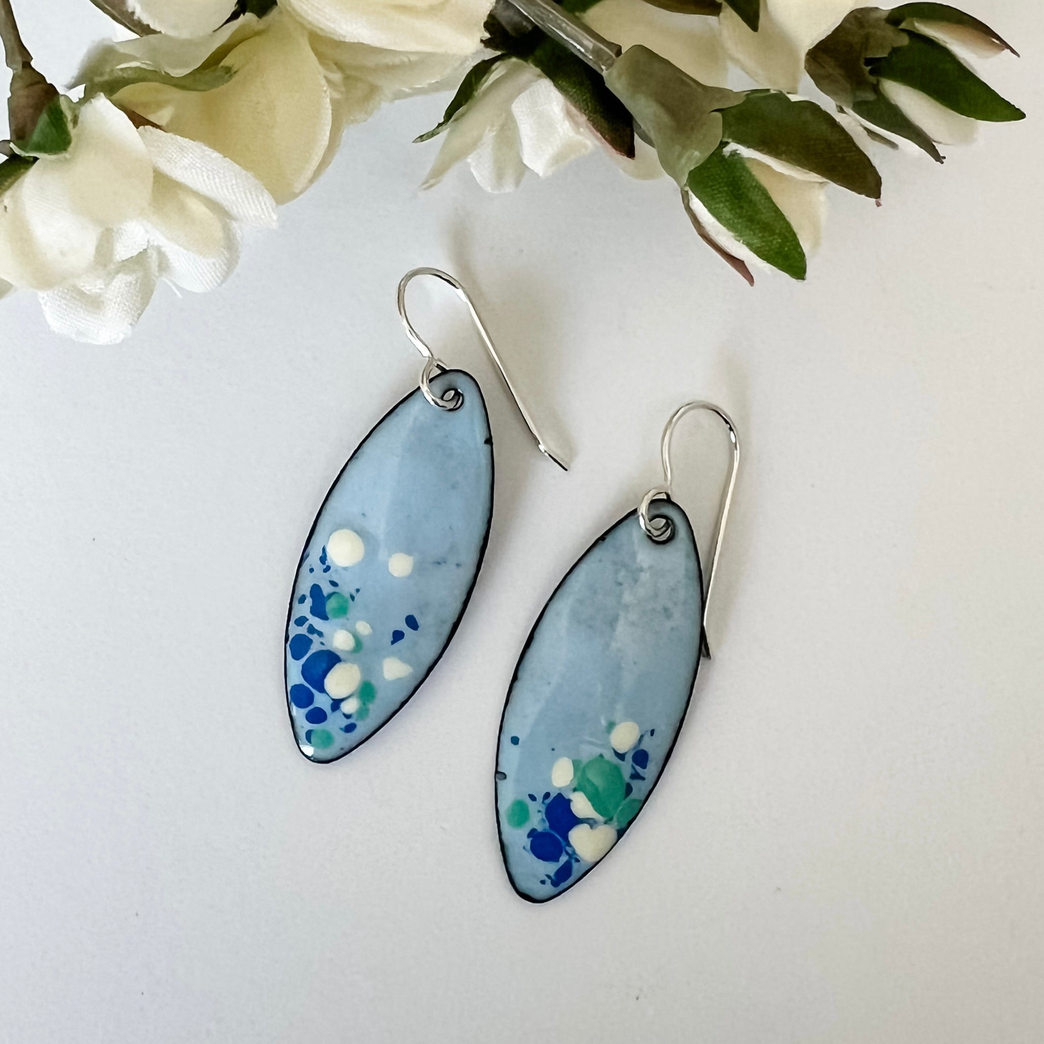 Handcrafted light blue enamel earrings with dark blue, cream, and seafoam splashes on sterling silver ear wires by Dawn Carlson Designs. Shown with fabric flowers.