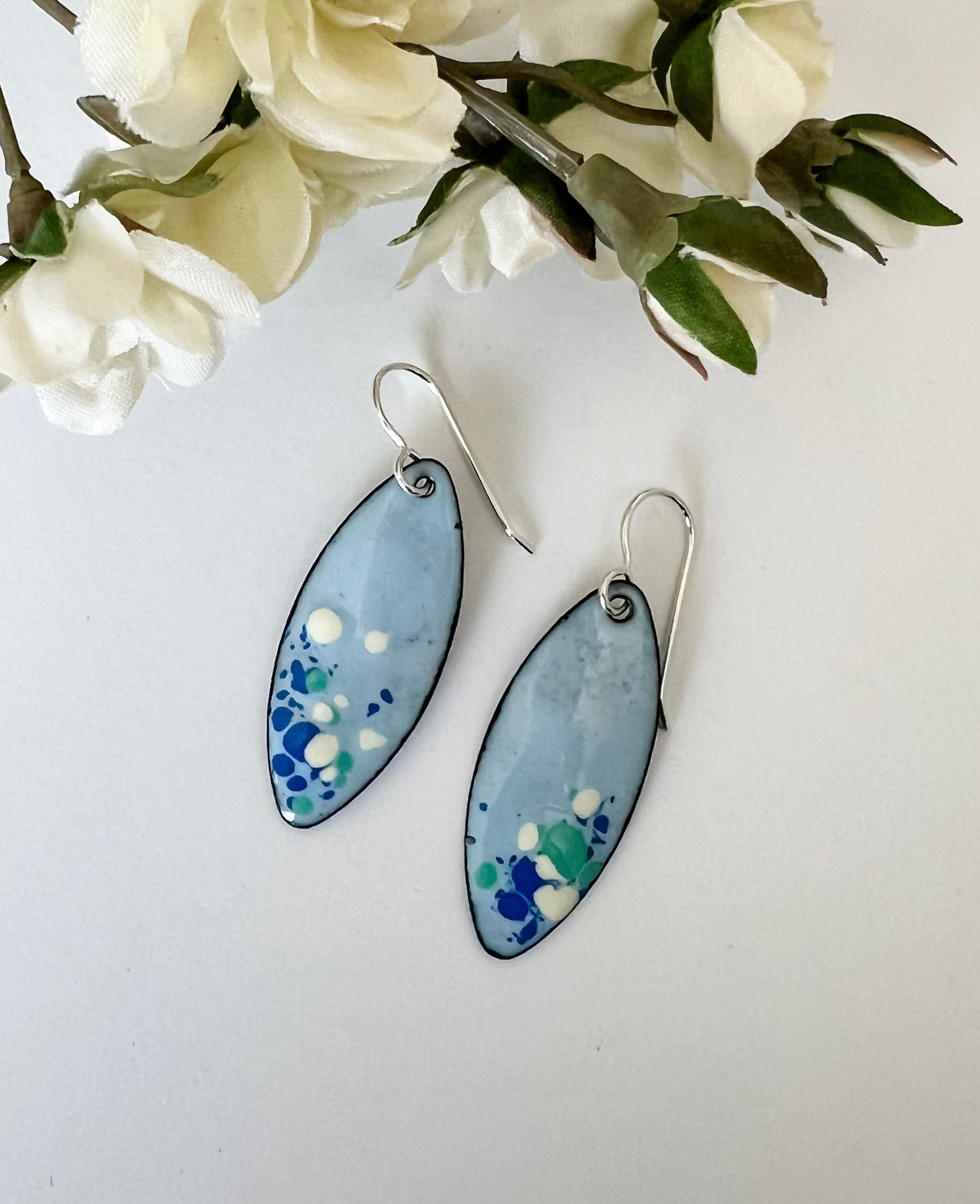 Handcrafted light blue enamel earrings with dark blue, cream, and seafoam splashes on sterling silver ear wires by Dawn Carlson Designs. Shown with fabric flowers.
