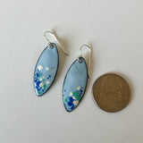 A pair of handmade enamel marquise drop earrings with light blue, green, and white speckled enamel and sterling silver fishhook wires, shown next to a US quarter for scale on a plain white surface.