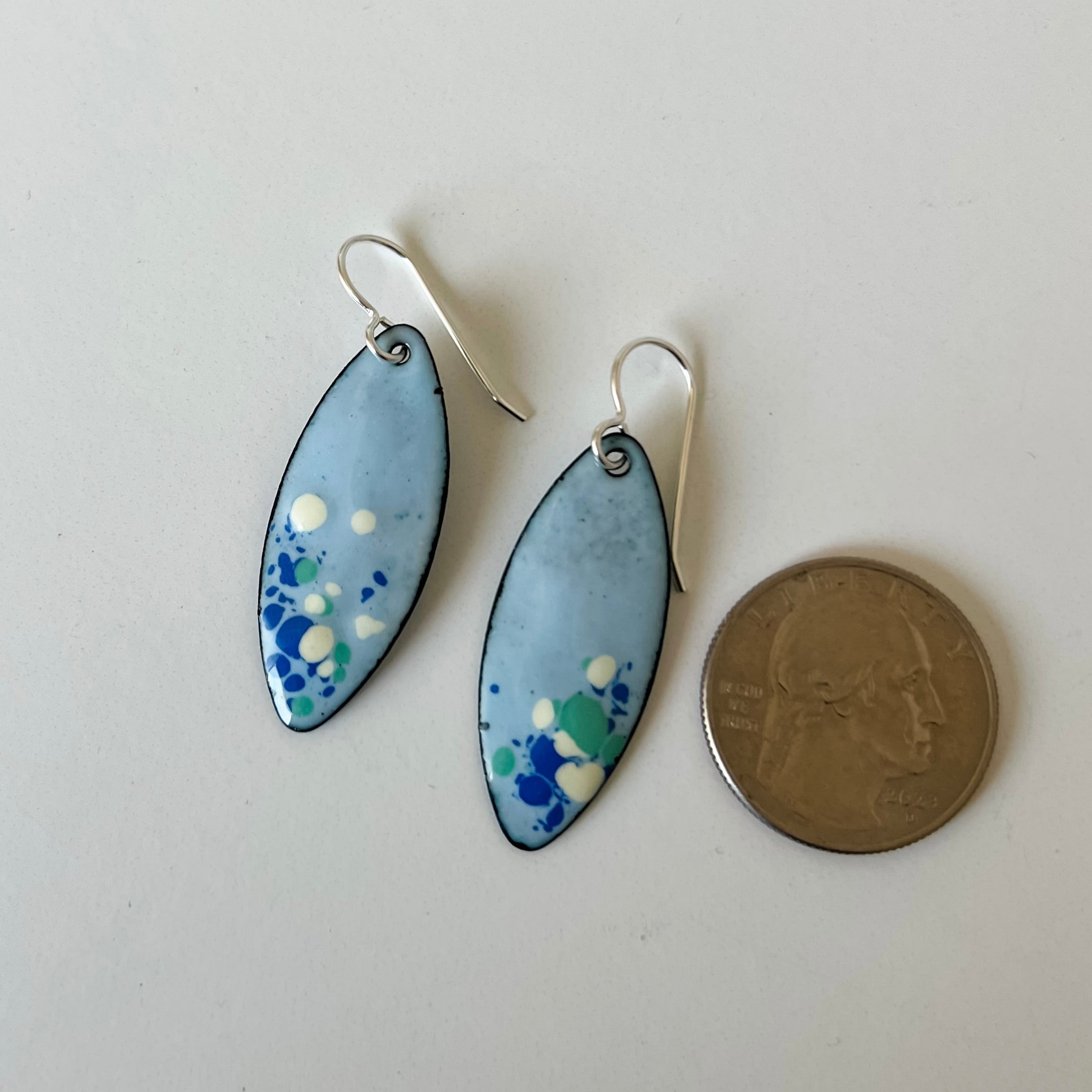 A pair of handmade enamel marquise drop earrings with light blue, green, and white speckled enamel and sterling silver fishhook wires, shown next to a US quarter for scale on a plain white surface.
