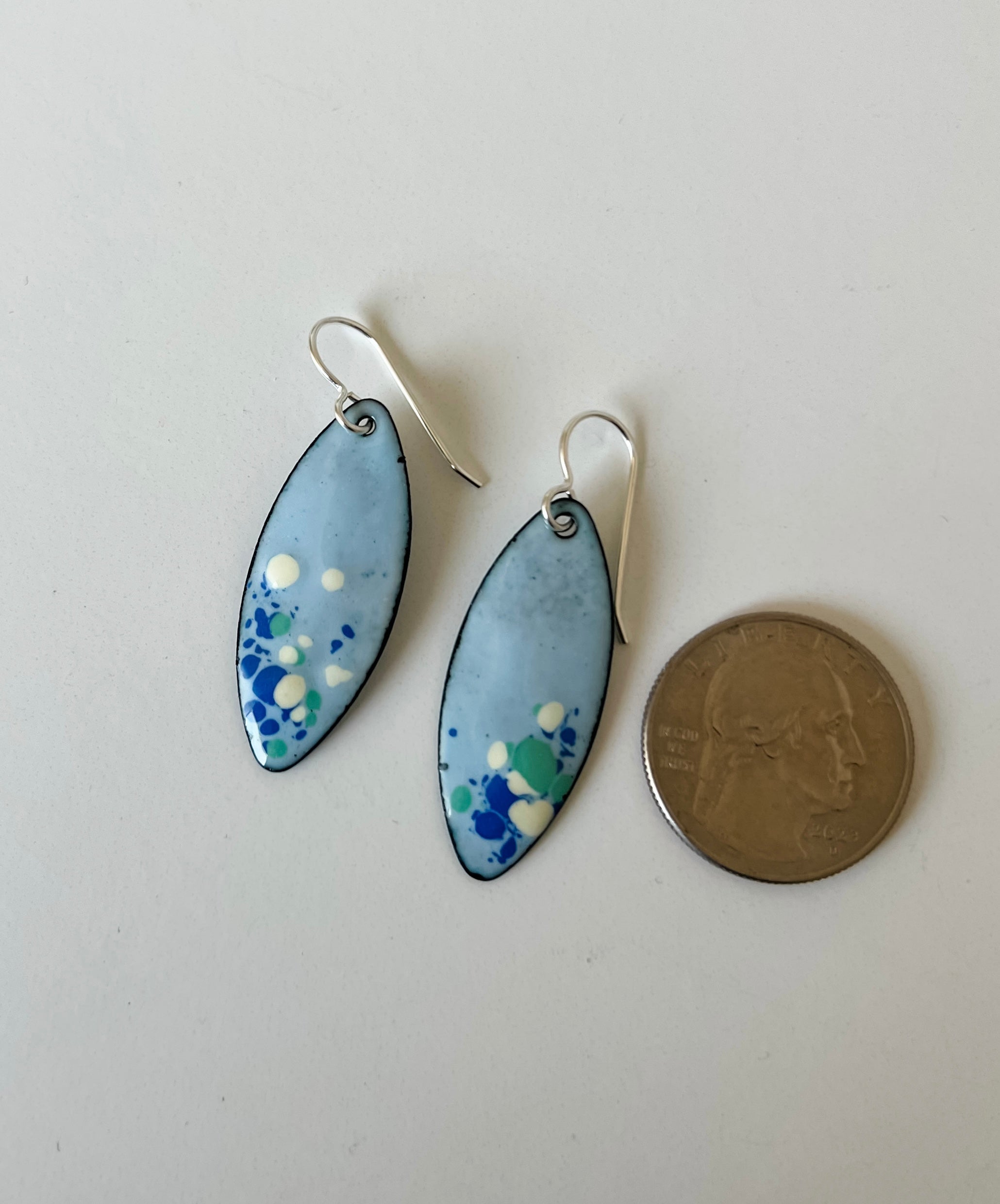A pair of handmade enamel marquise drop earrings with light blue, green, and white speckled enamel and sterling silver fishhook wires, shown next to a US quarter for scale on a plain white surface.