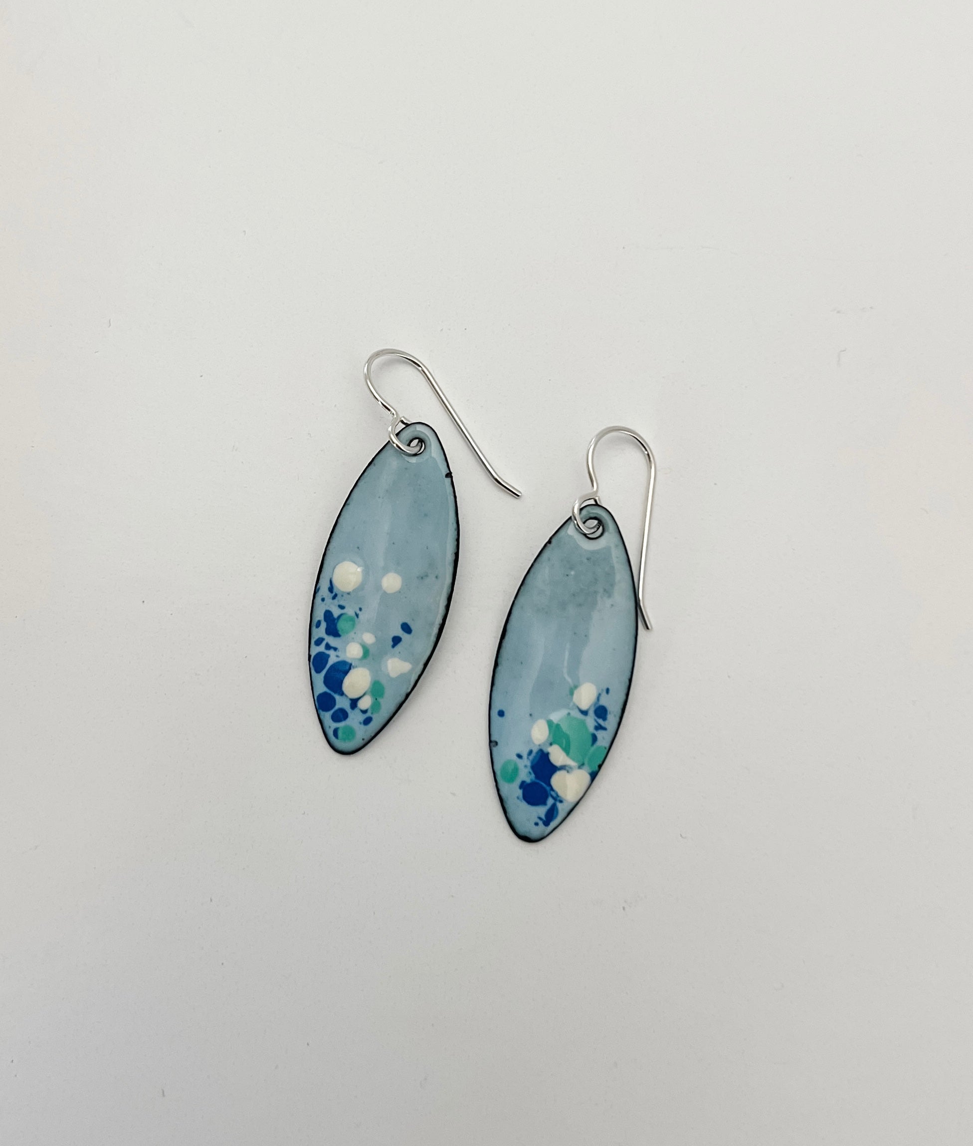 Handcrafted light blue enamel earrings with dark blue, cream, and seafoam splashes on sterling silver ear wires by Dawn Carlson Designs on a white background.
