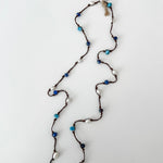 A lariat style necklace with dark brown cord, apatite, sodalite, white pearls, and a white branded tag at the top, laid out on a white background.