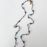 A lariat style necklace with dark brown cord, apatite, sodalite, white pearls, and a white branded tag at the top, laid out on a white background.