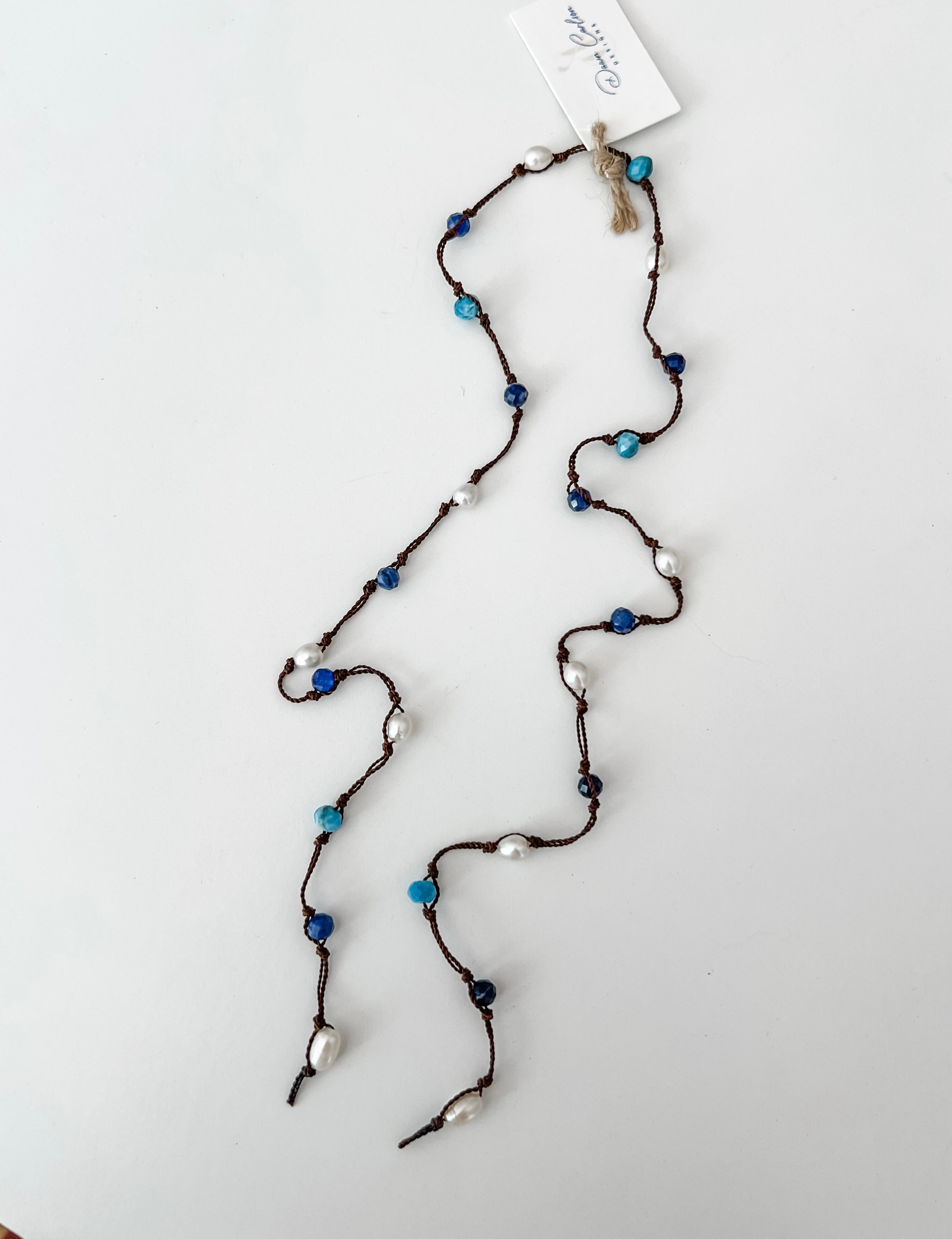 A lariat style necklace with dark brown cord, apatite, sodalite, white pearls, and a white branded tag at the top, laid out on a white background.